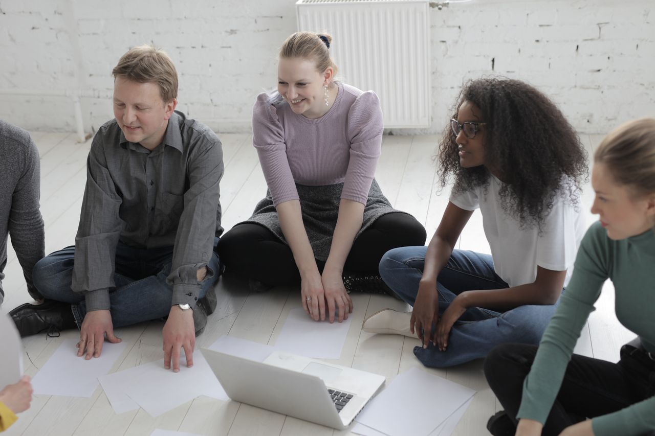 Glad colleagues sitting with crossed legs on ceramic floor near empty sheets of paper and open portable computer in afternoon behind brick wall